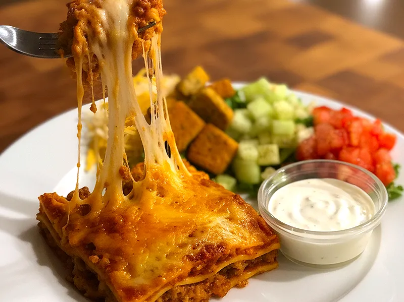 Cheesy baked lasagna with side salad fixings and ranch on a white plate